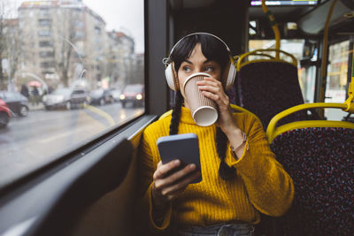 Woman wearing wireless headphones and drinking coffee in bus