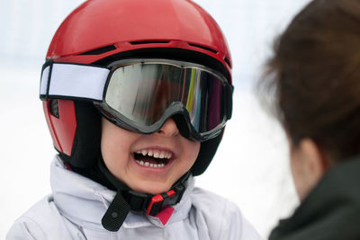 Boy in red helmet, ski goggles and white jacket smiling to his mother. winter sports, young skier