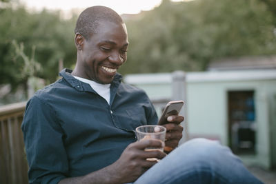 Smiling mid adult man using mobile phone while holding glass at porch