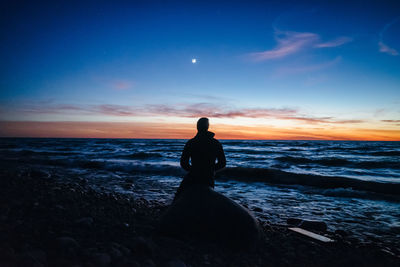 Man sitting on rock at beach against sky during sunset