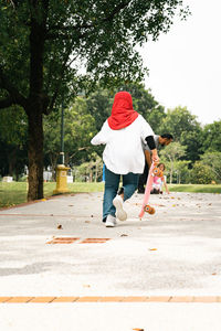 Young girl wearing hijab is running while holding pennyboard in the park. view from behind.