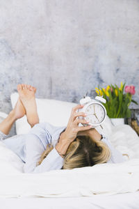 Woman with alarm clock lying on bed against wall at home