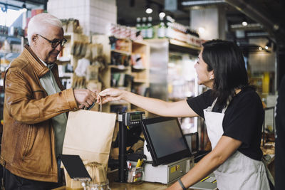 Smiling saleswoman giving shopping bag to senior male customer at store checkout