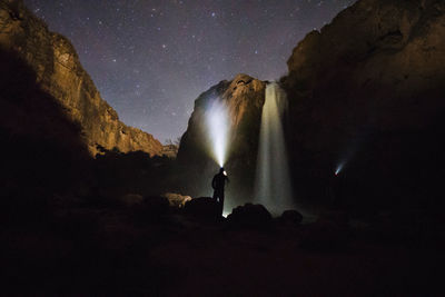 Silhouette man with flashlight standing by waterfall at night