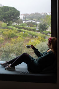 Side view of woman using mobile phone while sitting on window