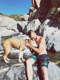 Shirtless young man petting dog while sitting on rock by waterfall during sunny day