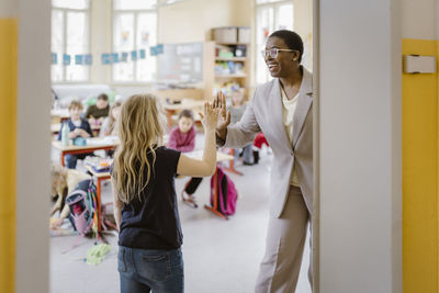 Smiling female teacher and schoolgirl giving high-five at doorway in classroom