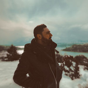 Young man looking away on snow against sky during winter