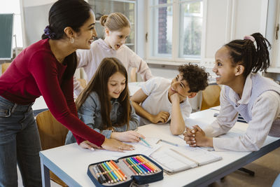 Happy female teacher leaning at desk with students in classroom