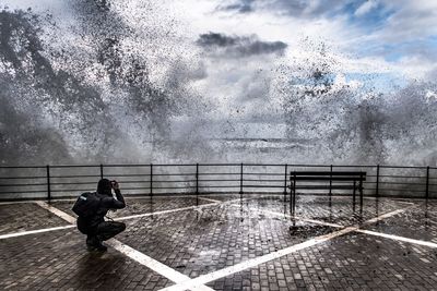 Man photographing splashing waves while crouching on promenade against sky