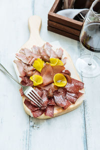 From above of slices of smoked meat different varieties creative served on wooden cutting board with metal fork and glass of red vine