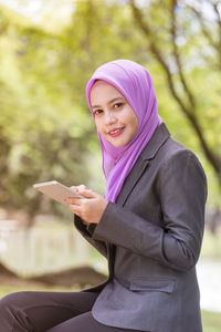 Portrait of young woman wearing hijab using mobile phone while sitting in park