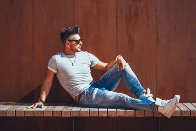 Young man looking away while sitting on wood against wall