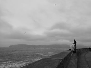 Men on retaining wall by sea against cloudy sky