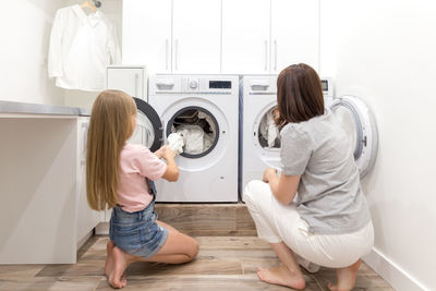 Rear view of mother and daughter pulling out clothes from washing machine at home