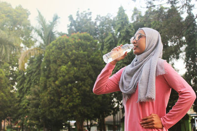 Young woman drinking water while standing against trees