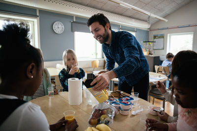 Teacher serving juice to students sitting at table in classroom