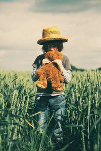 Boy holding teddy bear walking amidst wheat field against sky