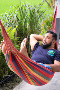 Asian bearded man is sitting in a hammock and looking at rice field and mountains .