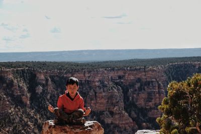 Boy meditating while sitting on rock formations against sky