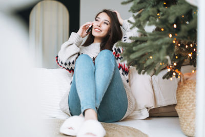 Young asian woman with long hair in cozy sweater using mobile on bed in room with christmas tree 