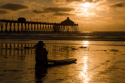 Silhouette woman sitting at beach against cloudy sky during sunset