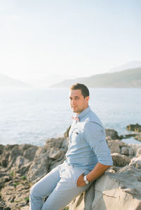 Portrait of young man sitting on rock by sea against clear sky