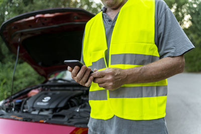 Close up of a senior man standing at his broken car wearing a safety vest and using his smartphone