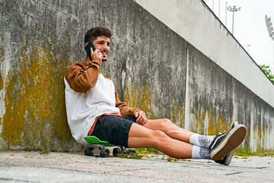 Young casual man sits outdoor on skateboard and uses phone person