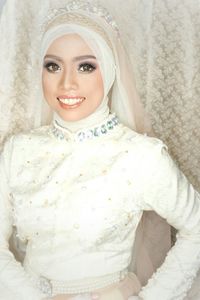 Portrait of smiling bride in hijab and white wedding dress standing against curtain