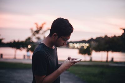 Side view of young man using mobile phone against lake at sunset