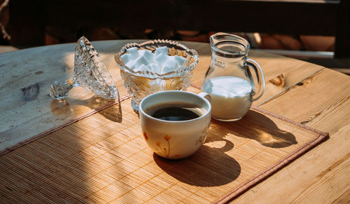 A cup of coffee stands outside on a table next to a jug of milk and a sugar bowl with lump sugar 