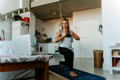 Concentrated young female in sportswear training with weight plate in kitchen at home watching tutorial on laptop