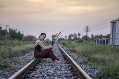 Portrait of woman holding plant while sitting on railroad track during sunset