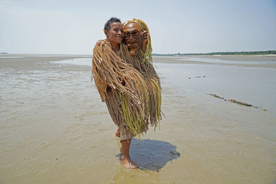 Portrait of mature man in costume holding mask while standing at beach against sky during sunny day