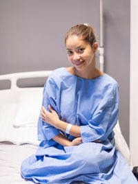 Young woman in a private hospital room, dressed in a blue gown, sits on a bed and waits to be