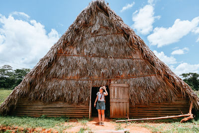 Resting lady in stylish casual clothing standing in doorway of big rural house with thatched roof and blue sky on background in cuba