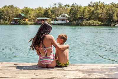 Rear view of mother and son sitting on pier over lake during sunny day