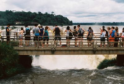 People standing on footbridge by waterfall against cloudy sky