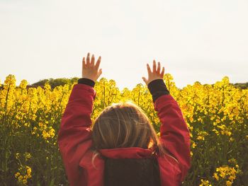 Rear view of girl with arms raised standing at oilseed rape against clear sky