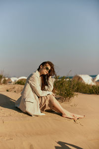 Young woman sitting on sand at desert against clear sky