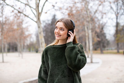 Smiling young woman listening music through headphones while looking away against bare trees during winter