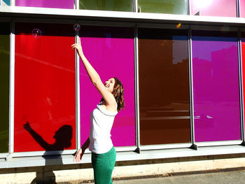Young woman reaching towards bubble while standing by colorful building