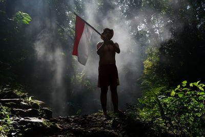 Full length of man holding flag standing against trees in forest