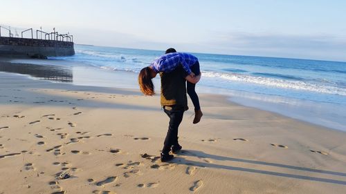 Rear view of playful man carrying girlfriend on shoulder at sandy beach