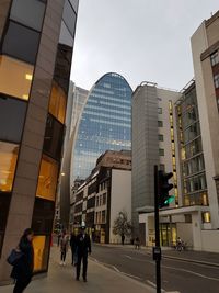 People walking on street by modern buildings in city against sky