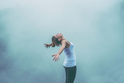 Side view of woman with arms outstretched standing in forest during foggy weather