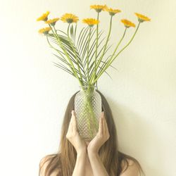 Woman holding flowers in vase in front of face against wall