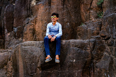 Low angle view of young man wearing sunglasses while sitting on rock