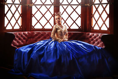 Woman wearing crown and evening gown sitting on sofa against window